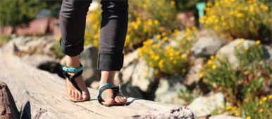 Eine Frau läuft über einen Holzsteg. Sie hat Luna Sandalen an. Zu sehen sind nur Füße und Unterschenkel.