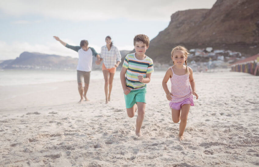 Am Meer: Zwei Kinder rennen lachend und barfuß über den Strand. Im Hintergrund gehen die Eltern spazieren.