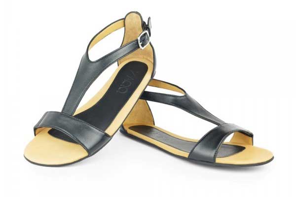 Die Barfuß-Sandal ZAQQ Chiq in schwarz mit einem schwarzen Lederriemen, der sich über Fußrücken, Zehen und Knöchel zieht und mit einer dezenten Metalllasche zugebunden wird.